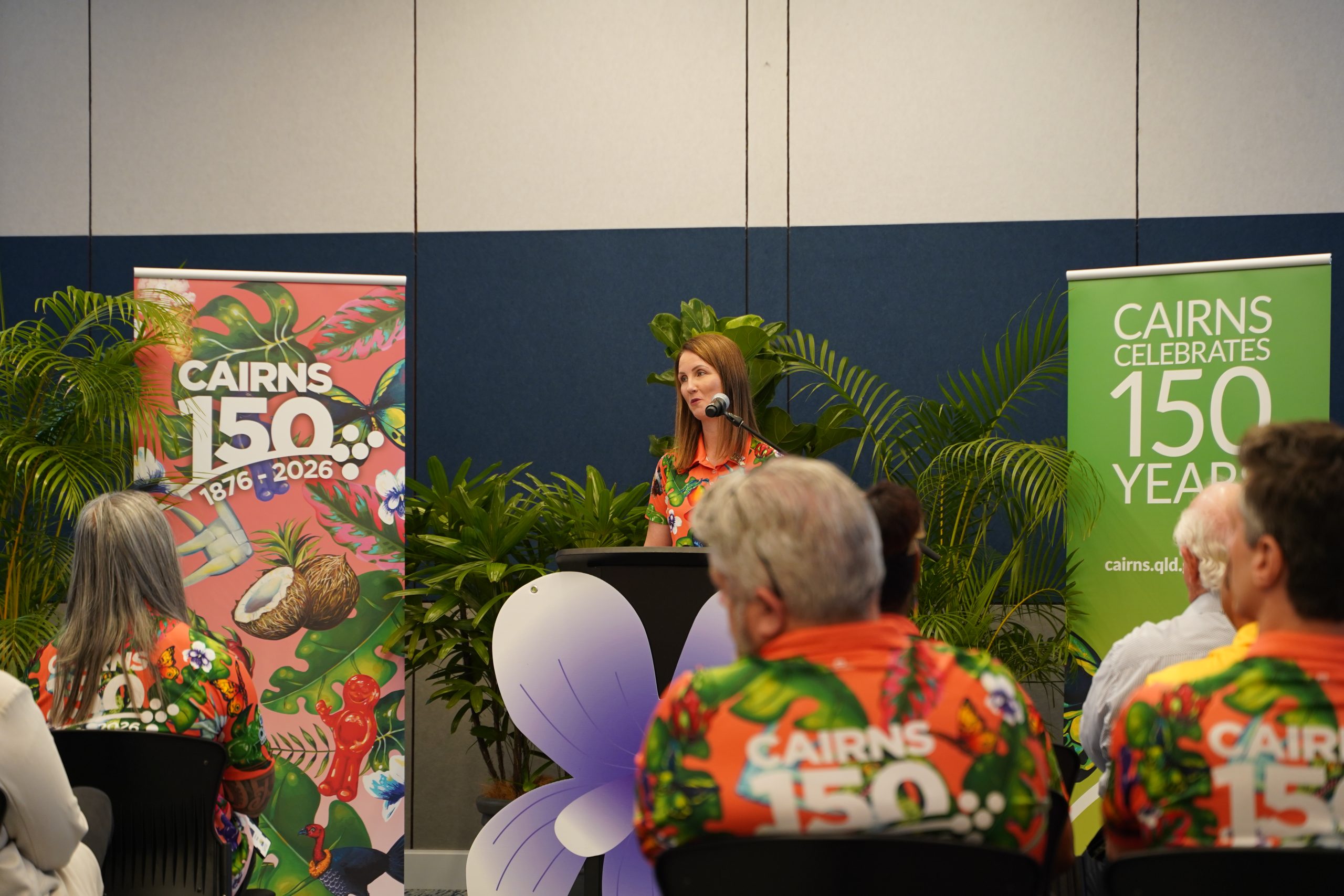Speaker at a podium during the Cairns 150 launch event, with Cairns 150 banners and audience members in the foreground